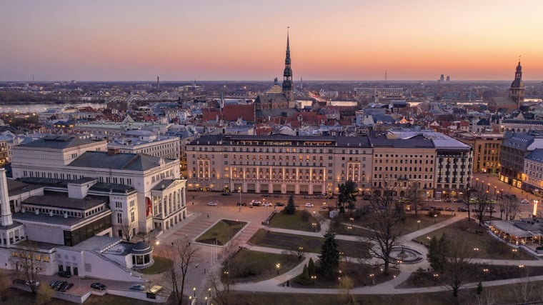 imagem 1Panoramic sunset view over Riga’s historic city center, featuring iconic buildings, tree-lined squares, and the city skyline illuminated at dusk.